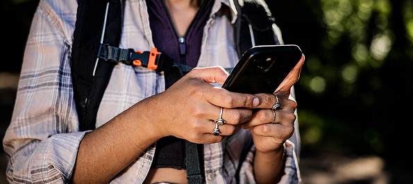 Hands holding a smartphone while a person wears a backpack and plaid shirt