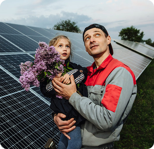 Man holding a child with lilac flowers, both looking up at the sky, in front of solar panels