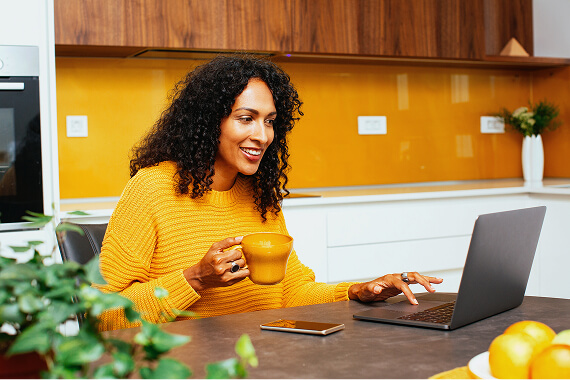 Smiling woman with curly hair in a yellow sweater using a laptop at a kitchen counter