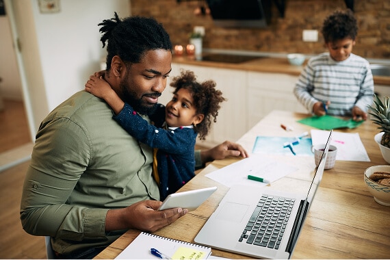 Father using a tablet with a child hugging him, another child crafts in the background