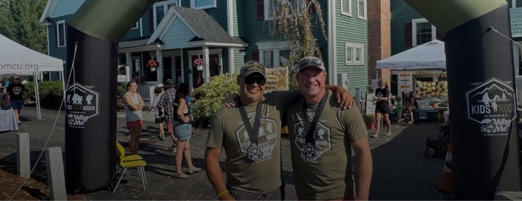 Two men wearing "Ruck March" medals and shirts smile with an event archway behind them