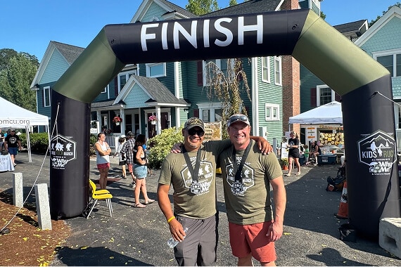 Two men wearing medals and matching Kids Ruck shirts smile under a finish line arch