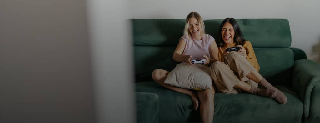 Two women laughing and playing video games on a green couch