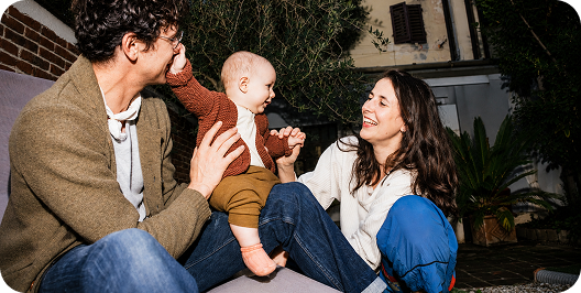 A happy family outdoors: a baby sits on an adult's lap, reaching for another adult's face, all smiling