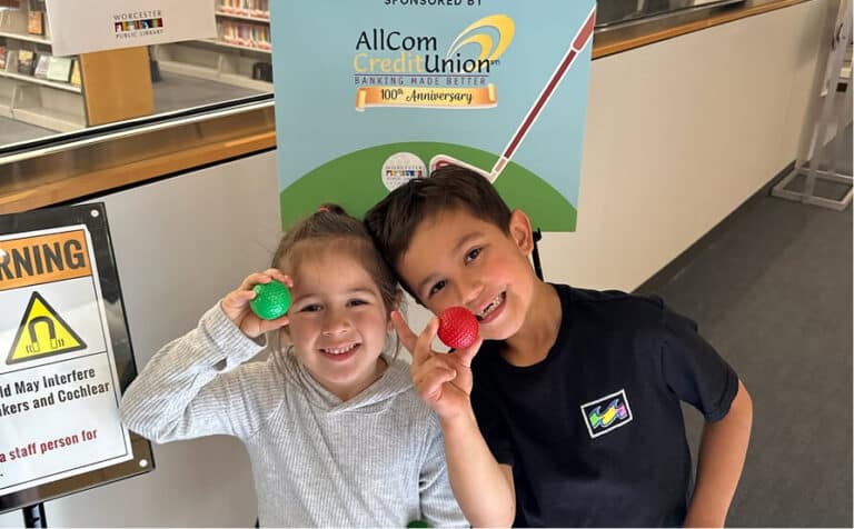Two smiling children holding green and red golf balls in front of an AllCom Credit Union banner