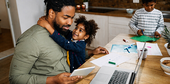 Father looks at a tablet while his child hugs him, with another child crafting nearby