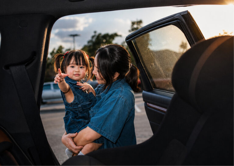 A woman holds a young child in denim overalls, viewed from inside a car with an open door
