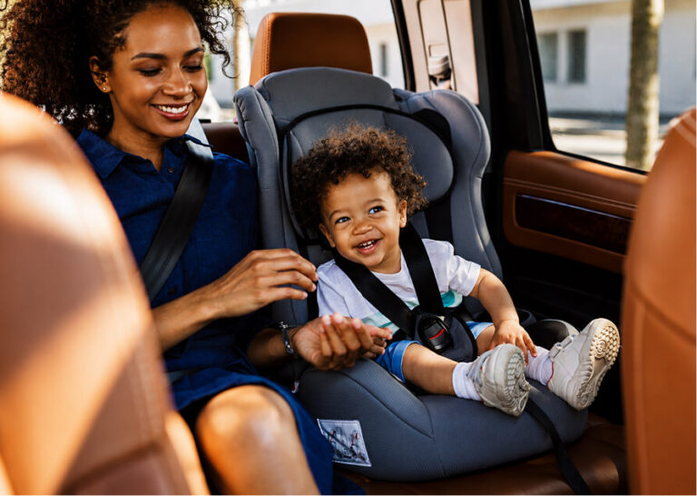 A smiling woman looks at a happy toddler secured in a car seat in the back of a car