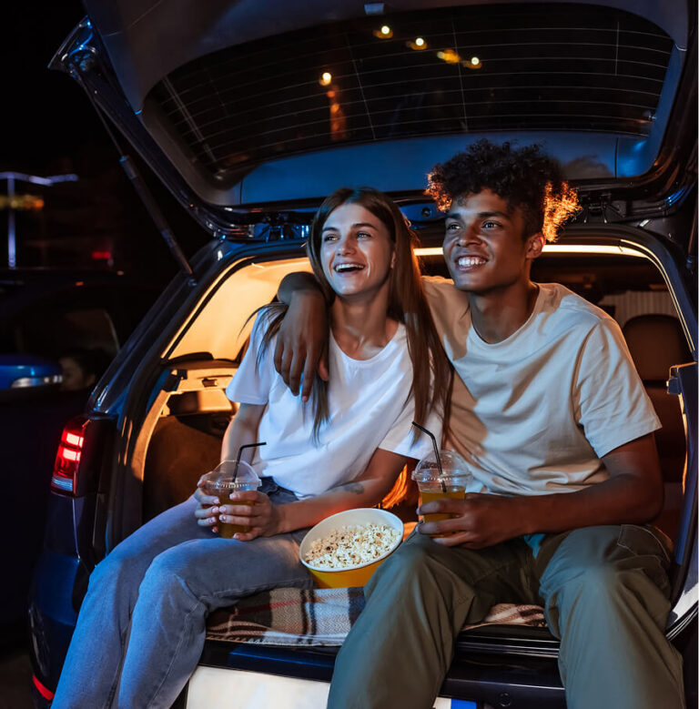 A young couple watching a drive-in movie from the trunk of their car, sharing popcorn and drinks