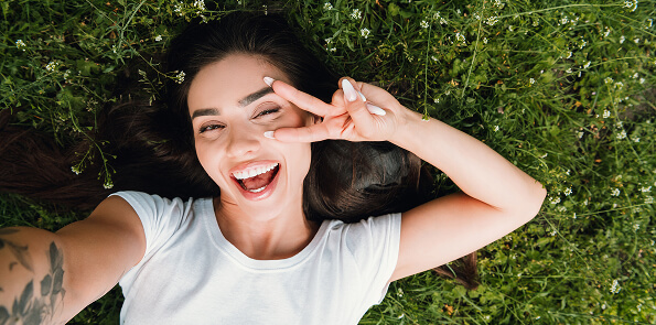 Smiling woman making a peace sign and laughing while lying in green grass