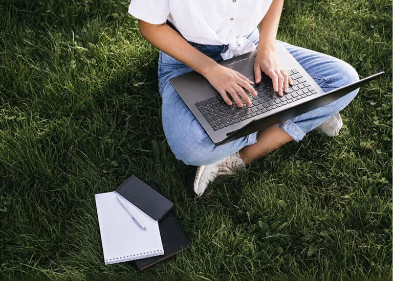 Person sitting on grass, typing on a laptop with a notebook and phone nearby