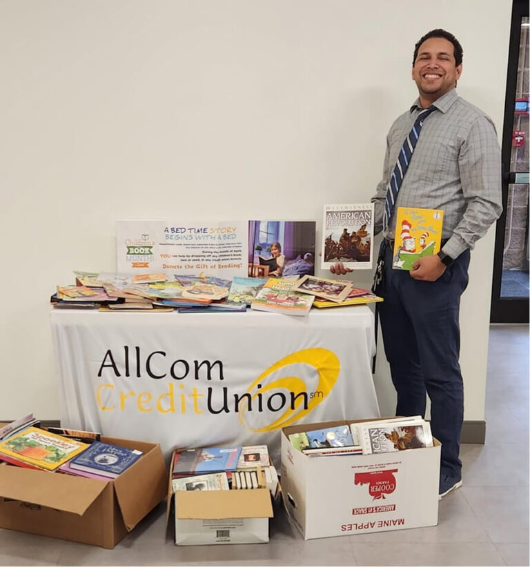 Man with two books standing next to a table filled with books and a “Donate the Gift of Reading” sign