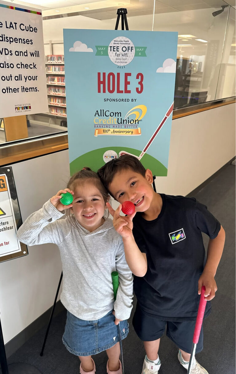 Two children holding golf balls and putters, smiling in front of a sign for a mini golf fundraiser