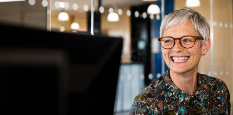 A smiling woman with short gray hair and glasses looks away from the camera, engaged and happy