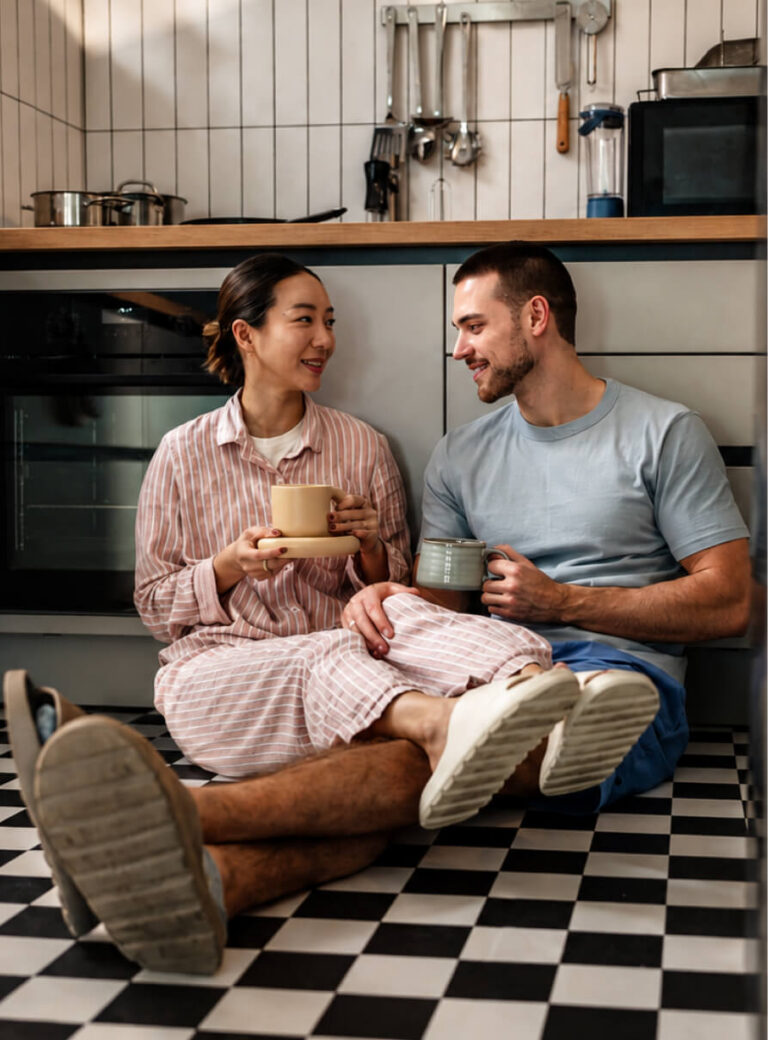 A couple sitting on a kitchen floor, drinking coffee and smiling at each other