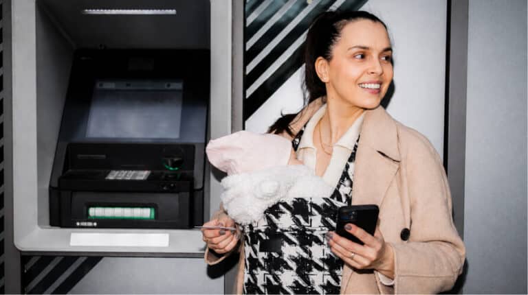 Woman with baby in carrier holding a credit card and phone next to an ATM