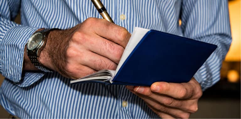 Person writing in a small blue notebook with a gold pen
