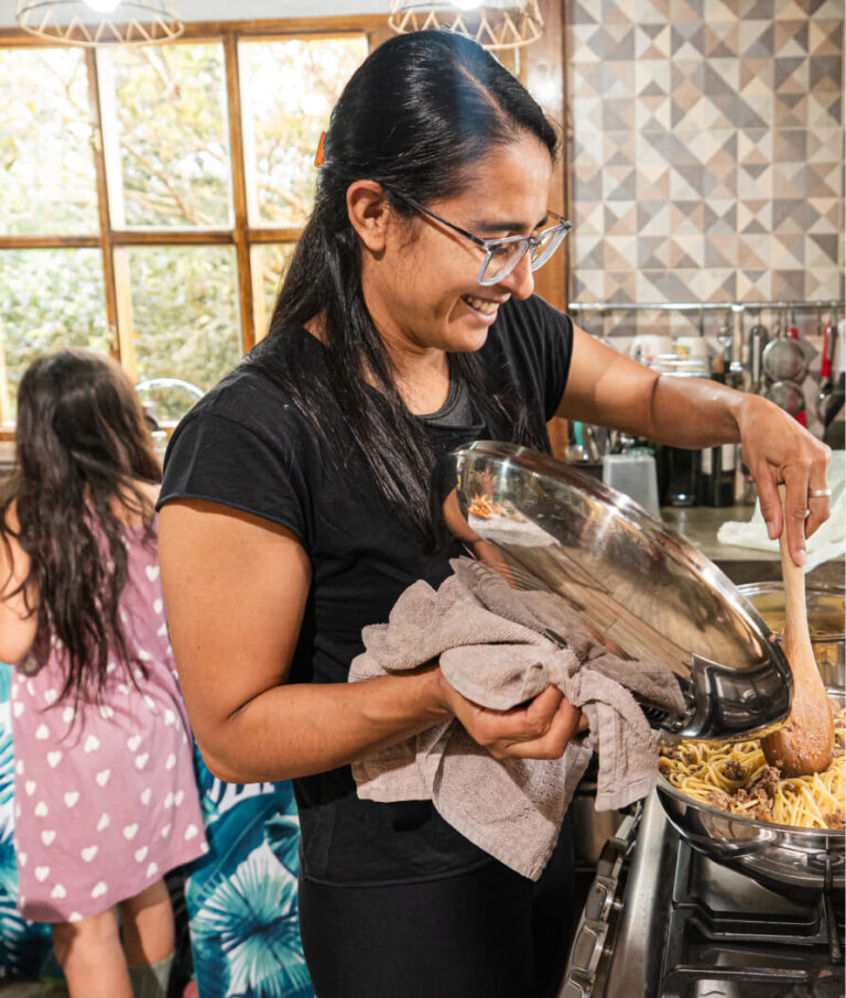 Woman stirring spaghetti with meat in a pan, holding a lid with a towel, smiling