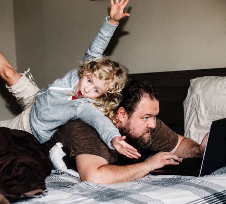 Child playing on a man's back while he uses a laptop on a bed