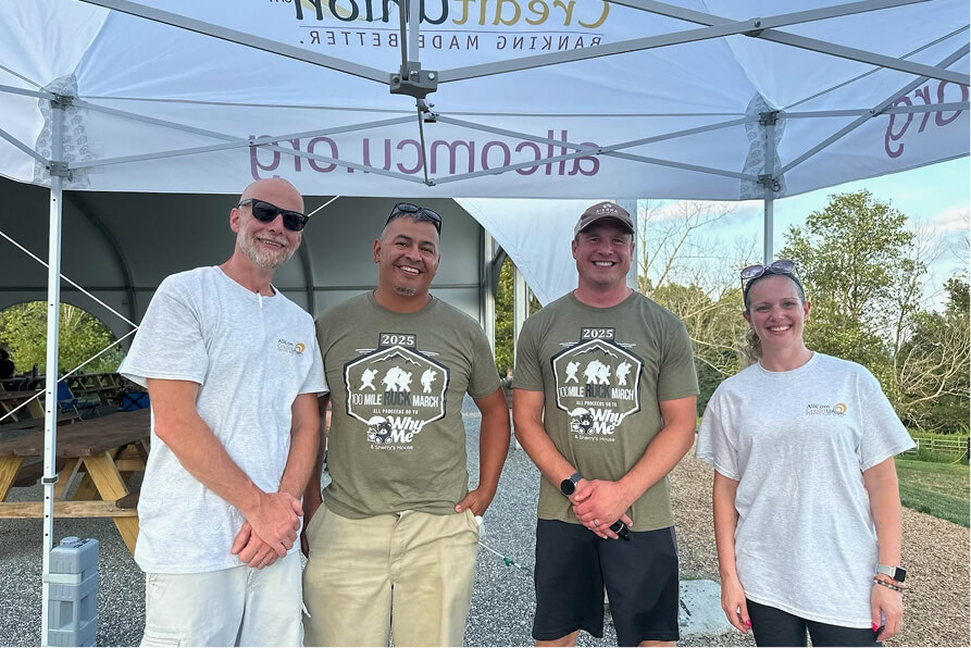 Four smiling people, two wearing "100 Mile Ruck March" shirts, standing under a tent