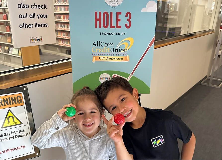 Two smiling children holding golf balls in front of a banner for a mini-golf fundraiser sponsored by AllCom Credit Union
