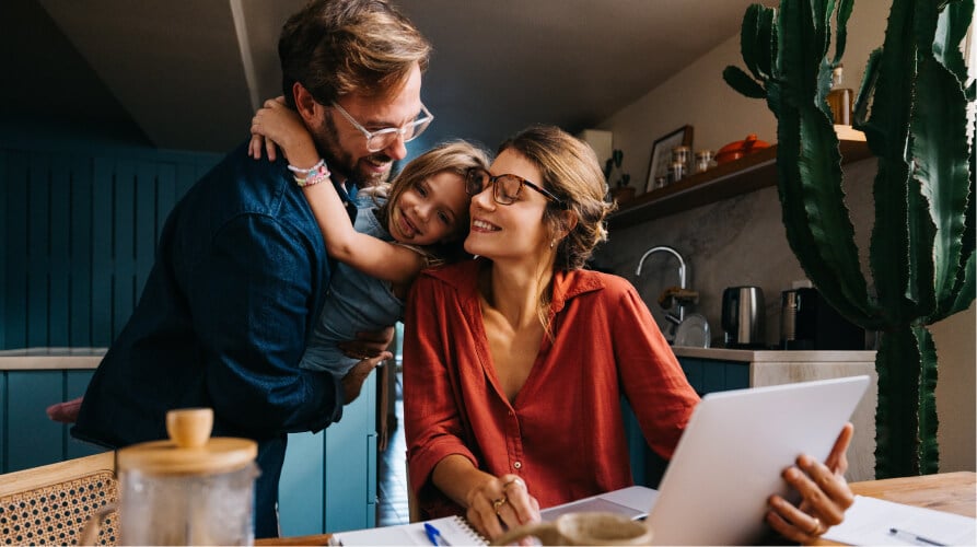A father holds his smiling child as they hug a mother who is working on a laptop at a kitchen counter
