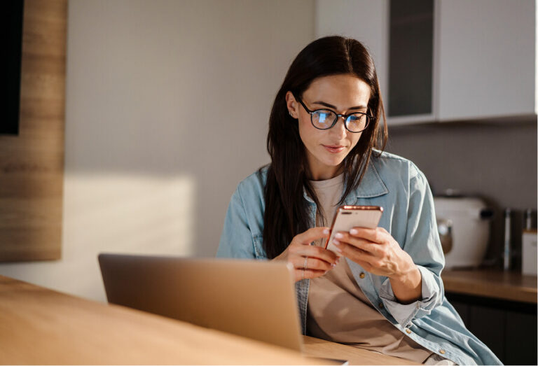 Woman wearing glasses looking at her phone, with a laptop open nearby