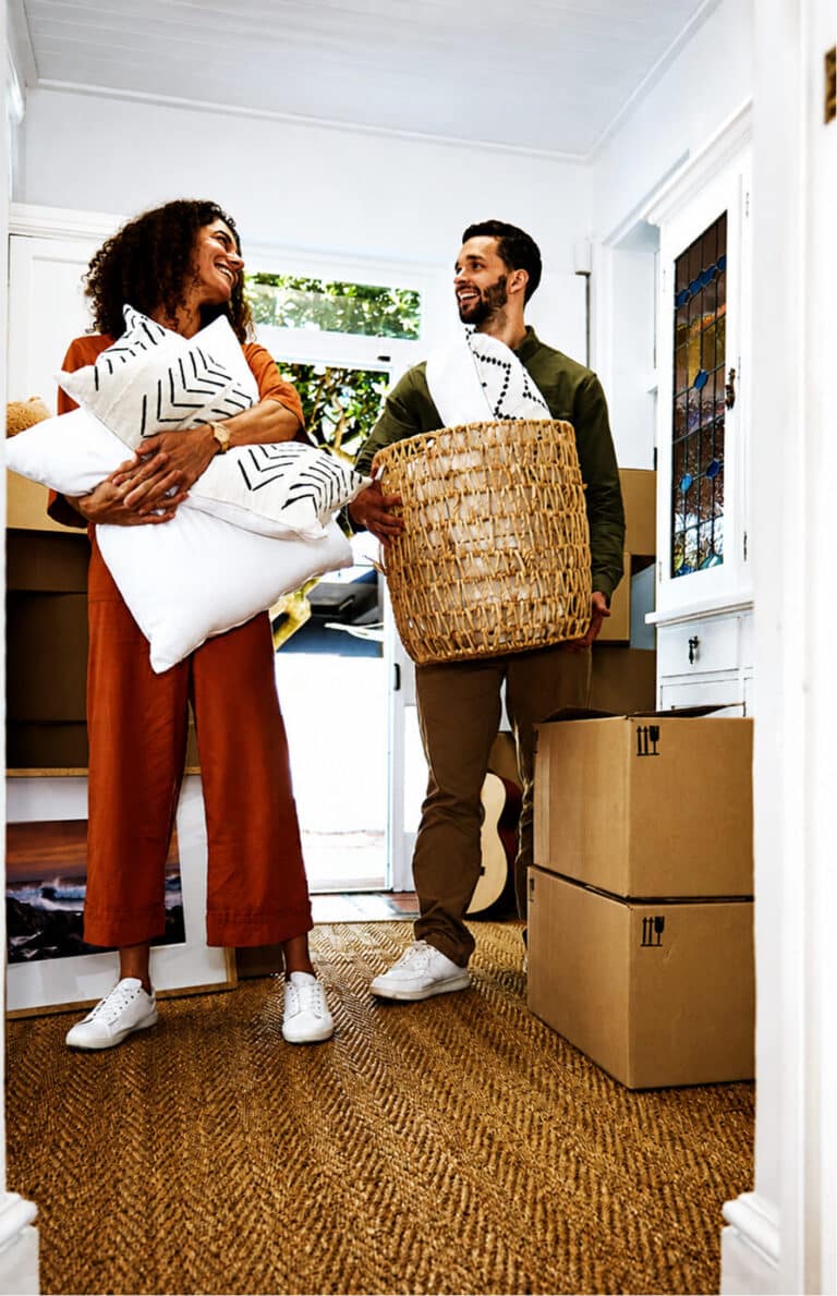A smiling couple moving into a new home, carrying pillows and a woven basket through an entryway with boxes.
