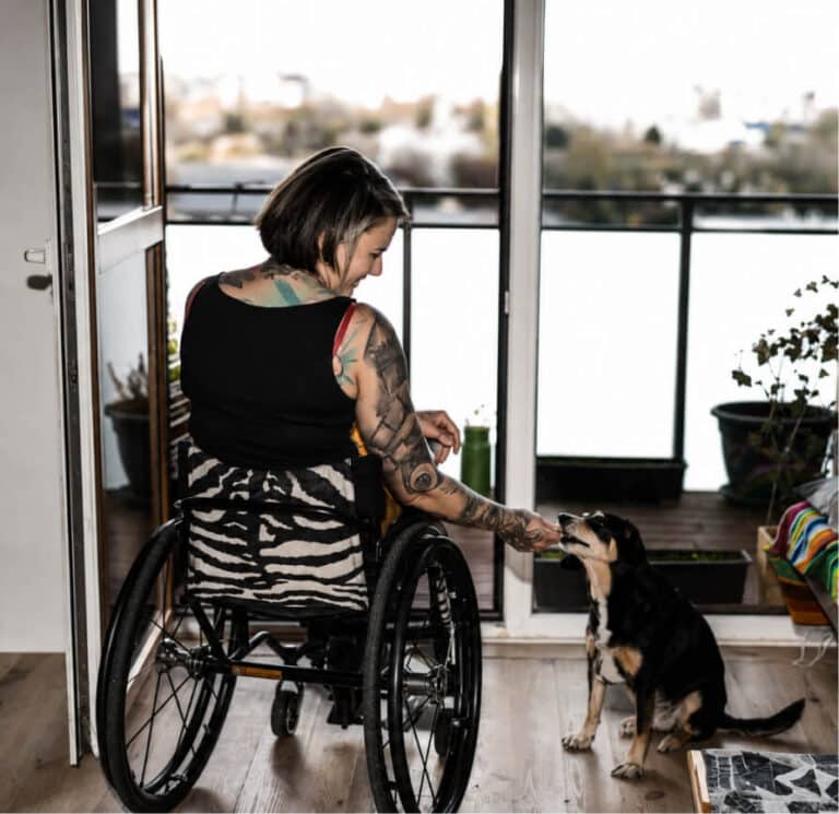Person in wheelchair feeding a small dog indoors, next to an open balcony door