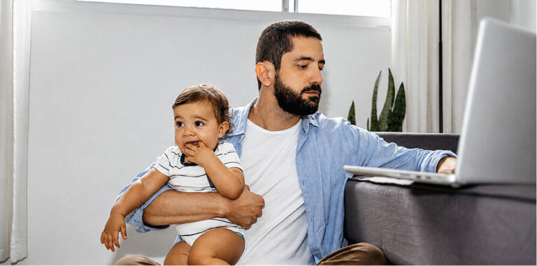 Man holding a baby while working on a laptop at home