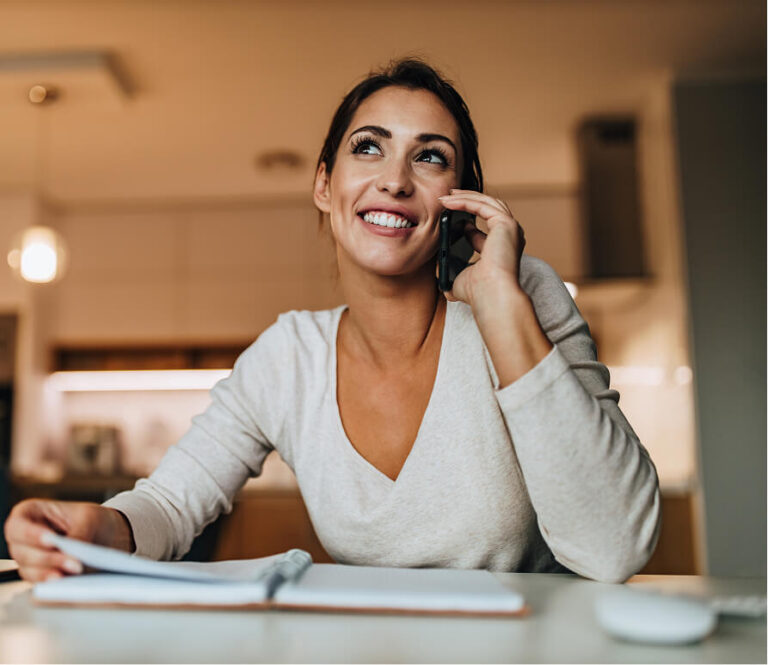 Smiling woman talking on a phone, looking upwards, while holding papers over a notebook