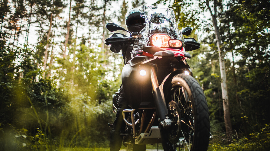 Person on an adventure motorcycle with headlights on, in a forest setting