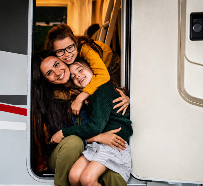 A woman and two young girls smiling and hugging in the doorway of a camper