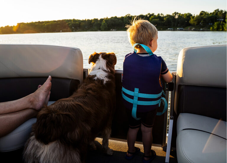 Child in a life vest and a dog looking out from a boat over a lake at sunset