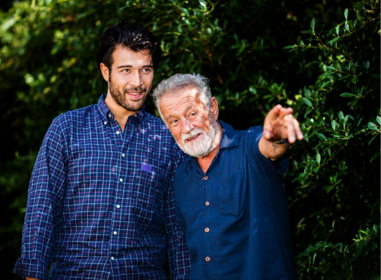 Older man with white beard smiles and points while younger man listens beside him