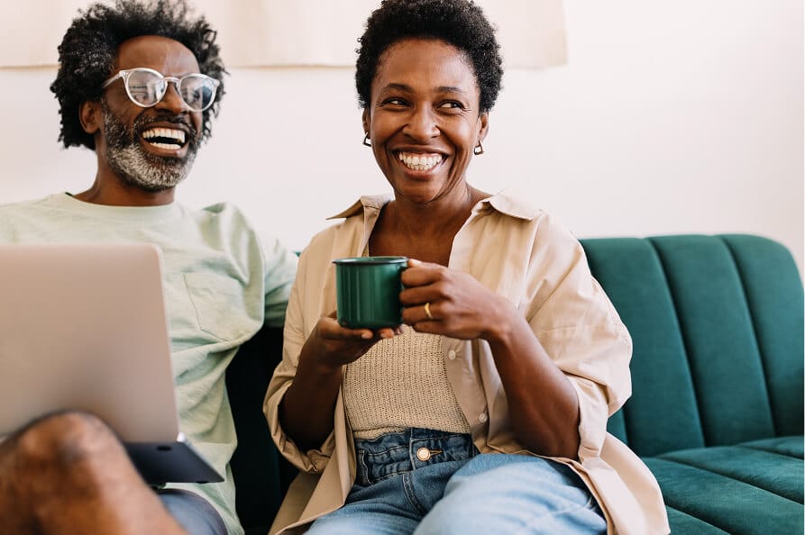 Two people smiling and laughing while relaxing, one with a laptop, the other with a mug