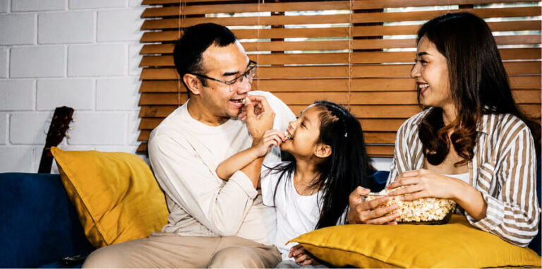 Father, mother, and child sharing popcorn on a couch, smiling and laughing together