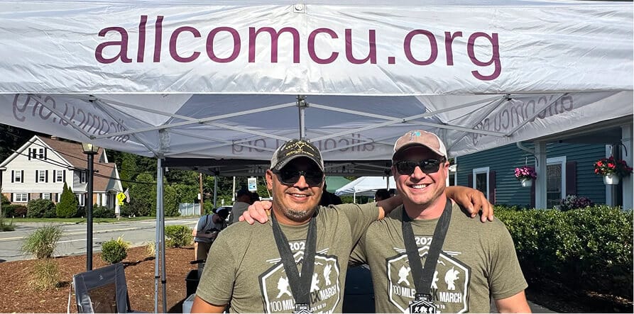 Two men smiling under a tent displaying "allcomcu.org", wearing shirts and medals from a "100 Mile Ruck March"