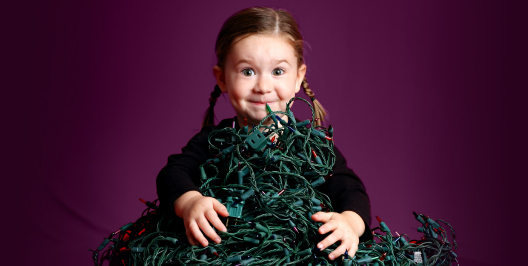 Young girl with pigtails smiling behind a large tangled pile of green Christmas lights